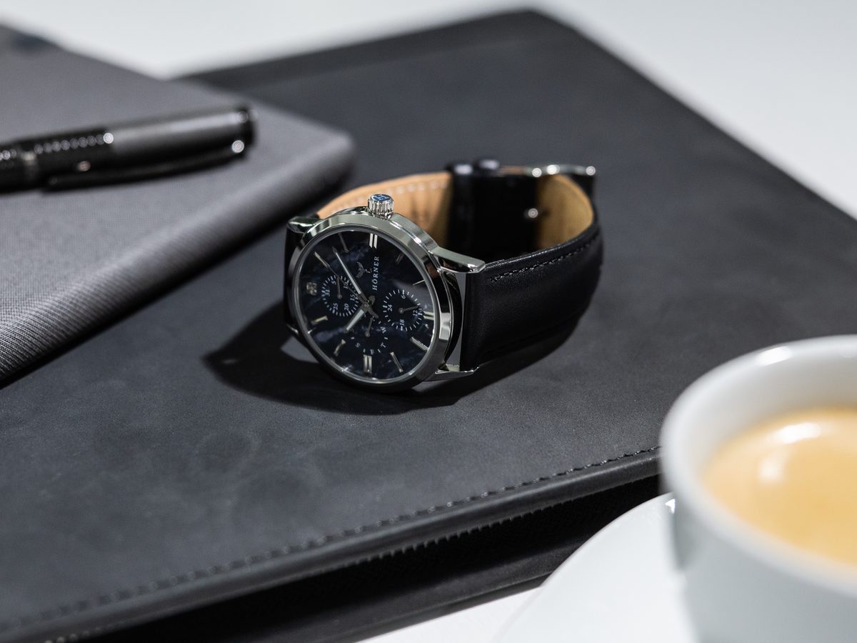 Armbanduhr mit mitternachtsblauem Zifferblatt und schwarzem Lederband auf einer Schreibmappe, daneben ein Schreibgerät und eine Kaffeetasse