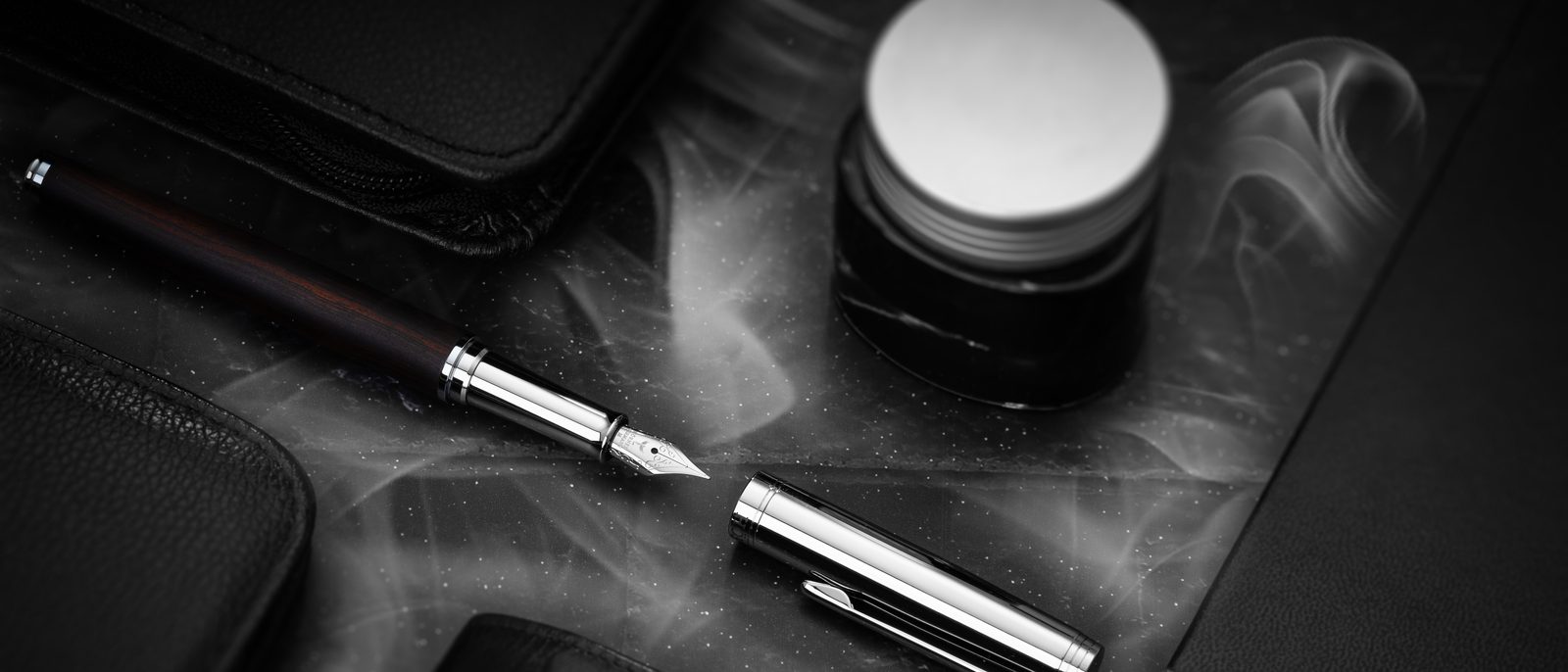 Un stylo-plume Hörner Legno en bois sombre avec sa plume visible, à côté d’un flacon d’encre ouvert et d’étuis en cuir sur un fond sombre
