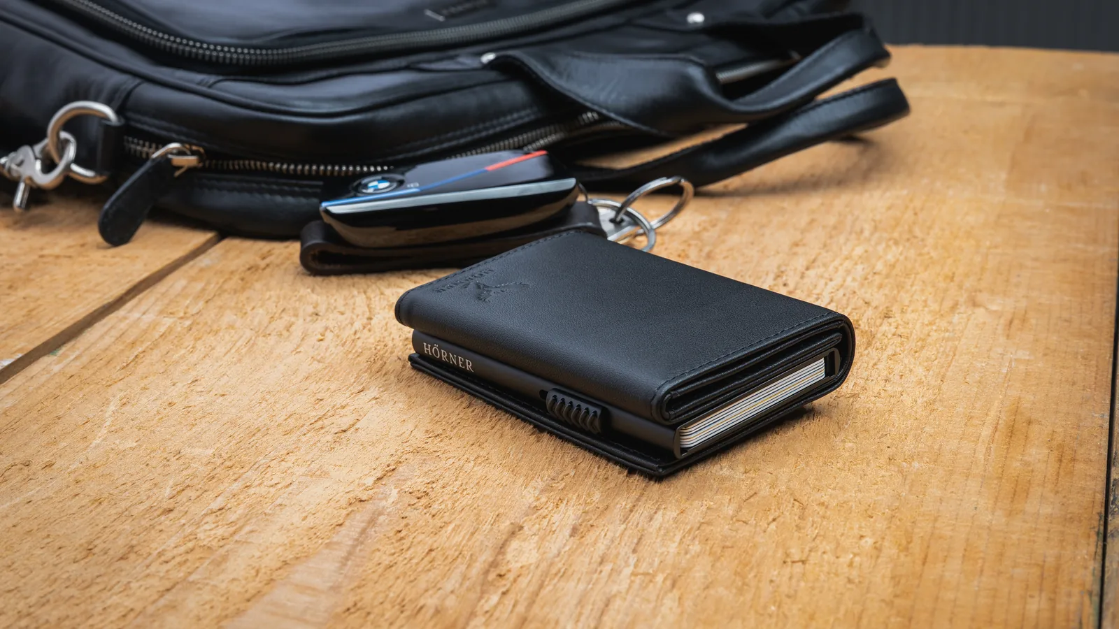 Slim Wallet Hörner Nexus noir posé sur une table en bois, à côté d’une pochette en cuir et d’une clé de voiture, un porte-cartes Pop-Up avec mécanisme Easy-Slide
