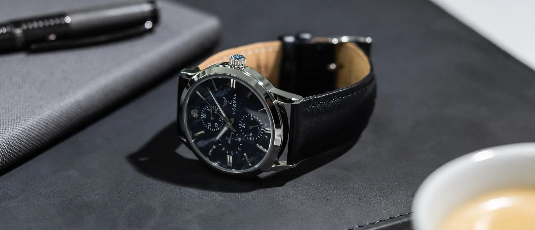Hörner Nova Midnight, eine Armbanduhr mit dunkelblauem Zifferblatt in Perlmutt-Optik, schwarzem Lederband und poliertem Stahlgehäuse, liegt auf einer schwarzen Mappe; links oben ein dunkler Stift auf einem grauen Notizbuch, rechts unten unscharf eine Tasse mit Espresso